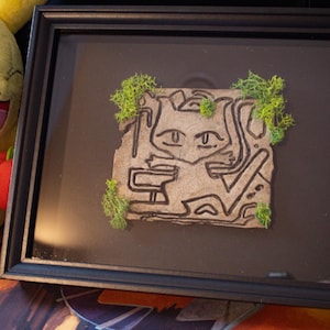 Puede incluir: Obra de arte enmarcada que presenta una ilustración estilizada de un personaje de dibujos animados. La obra de arte está sobre un fondo texturizado de color marrón claro, con acentos de musgo verde. El marco es negro. Dos peluches de Pikachu flanquean la obra.