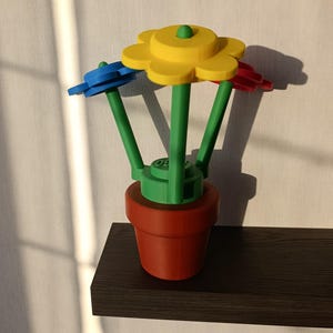 Op de afbeelding: Een 3D-geprinte bloempot met drie bloemen in primaire kleuren. De bloemen zijn geel, blauw en rood, met groene stelen en een groene basis. De pot is roodbruin en staat op een donkerbruin plankje.