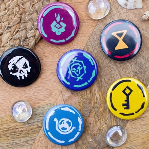 May include: Six round, colorful buttons with different symbols on them. The symbols include a skull, a key, an hourglass, a flame, a spiral, and a circle with a dot in the center. The buttons are on a wooden surface.