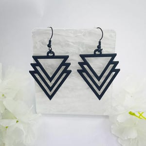 May include: A pair of black geometric earrings. Each earring has a layered triangle design suspended from a black hook. The earrings are displayed on a white, textured surface, with white flowers in the background.