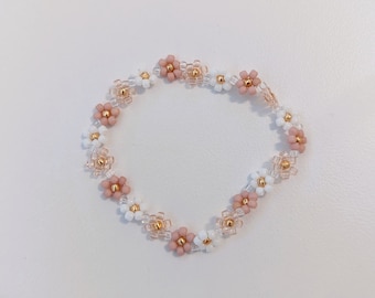 May include: A beaded bracelet featuring a floral design. The bracelet alternates between white and dusty rose-colored flower beads, with gold-colored beads at the center of each flower. The word "Babe" is in the upper right corner.