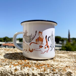 Op de afbeelding: Een witte emaille mok met een zwarte rand en handvat, voorzien van een kleurrijke illustratie van diverse katten in verschillende houdingen, waaronder een grijze cyperse kat, een oranje kat, een lapjeskat en een grijze kat rondom de beker.