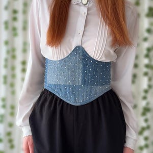 Könnte beinhalten: Ein Jeans-Korsett mit weißen Tupfen und Zierleiste. Das Korsett wird über einem weißen Hemd mit Kragen und einer schwarzen Hose getragen. Eine Vintage-Brosche ist am Hemdkragen befestigt. Das Korsett ist ein Taillengürtel.