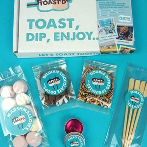 May include: A marshmallow roasting kit on a blue background. The kit includes a small, silver, round, micro toaster, a bag of white and pink marshmallows, a bag of chocolate chips, a bag of sprinkles, and four wooden skewers.