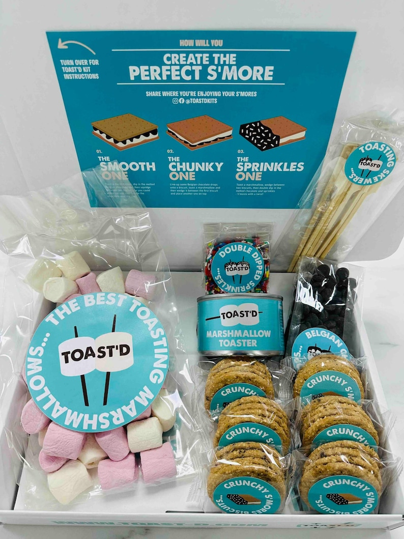 May include: A white box filled with ingredients for making s'mores. The box includes a can of marshmallows, a bag of chocolate chips, a bag of graham crackers, and a bag of sprinkles. The box also includes a small wooden stick for roasting marshmallows.