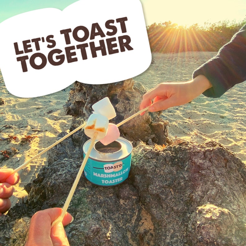 May include: A blue and white marshmallow toaster with the text "Toast'd Marshmallow Toaster" on the side. Two people are roasting marshmallows on sticks over the toaster. The text "Let's Toast Together" is in a white speech bubble in the top left corner of the image.