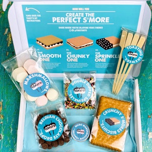 May include: A white box with a blue lid containing a s'mores kit. The kit includes marshmallows, chocolate chips, graham crackers, sprinkles, and skewers. The box has the text "Create the Perfect S'more" on the lid.