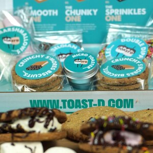 May include: A close-up shot of a s'mores kit with various components. The kit includes graham crackers, marshmallows, and chocolate. The packaging features the text "Create the Perfect S'More" and "Crunchy S'mores Biscuits". The website www.toast-d.com is also visible.