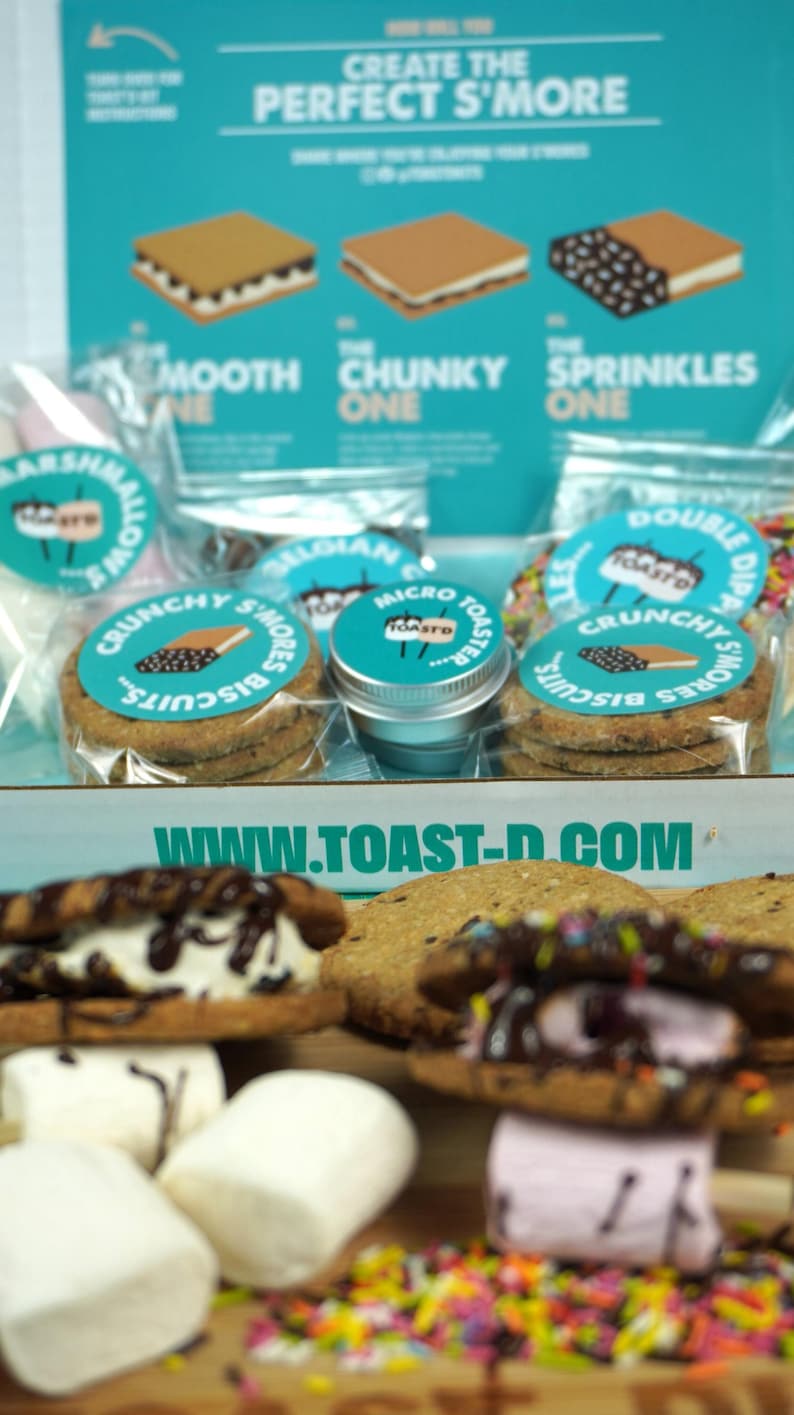 May include: A close-up shot of a s'mores kit with various components. The kit includes graham crackers, marshmallows, and chocolate. The packaging features the text "Create the Perfect S'More" and "Crunchy S'mores Biscuits". The website www.toast-d.com is also visible.