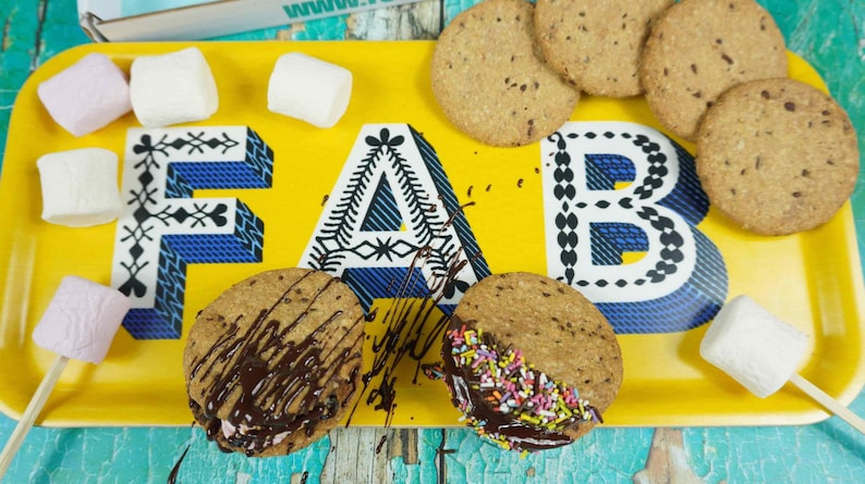 May include: A yellow tray with the word "FAB" in blue and white letters holds cookies and marshmallow treats. Some cookies are drizzled with chocolate and sprinkles. Several marshmallow pops are arranged around the tray.