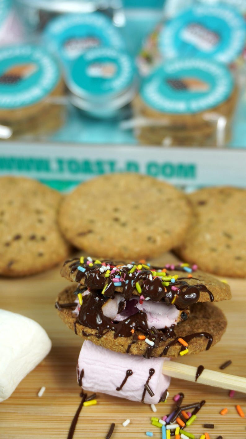 May include: A close-up of a s'more on a wooden skewer, featuring two round cookies with chocolate drizzle and colorful sprinkles, and a pink marshmallow. Several cookies and packaged s'mores are in the background.