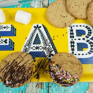 May include: A yellow tray with the word "FAB" printed on it. The tray is filled with cookies, marshmallows, and chocolate. The cookies are topped with chocolate and sprinkles.