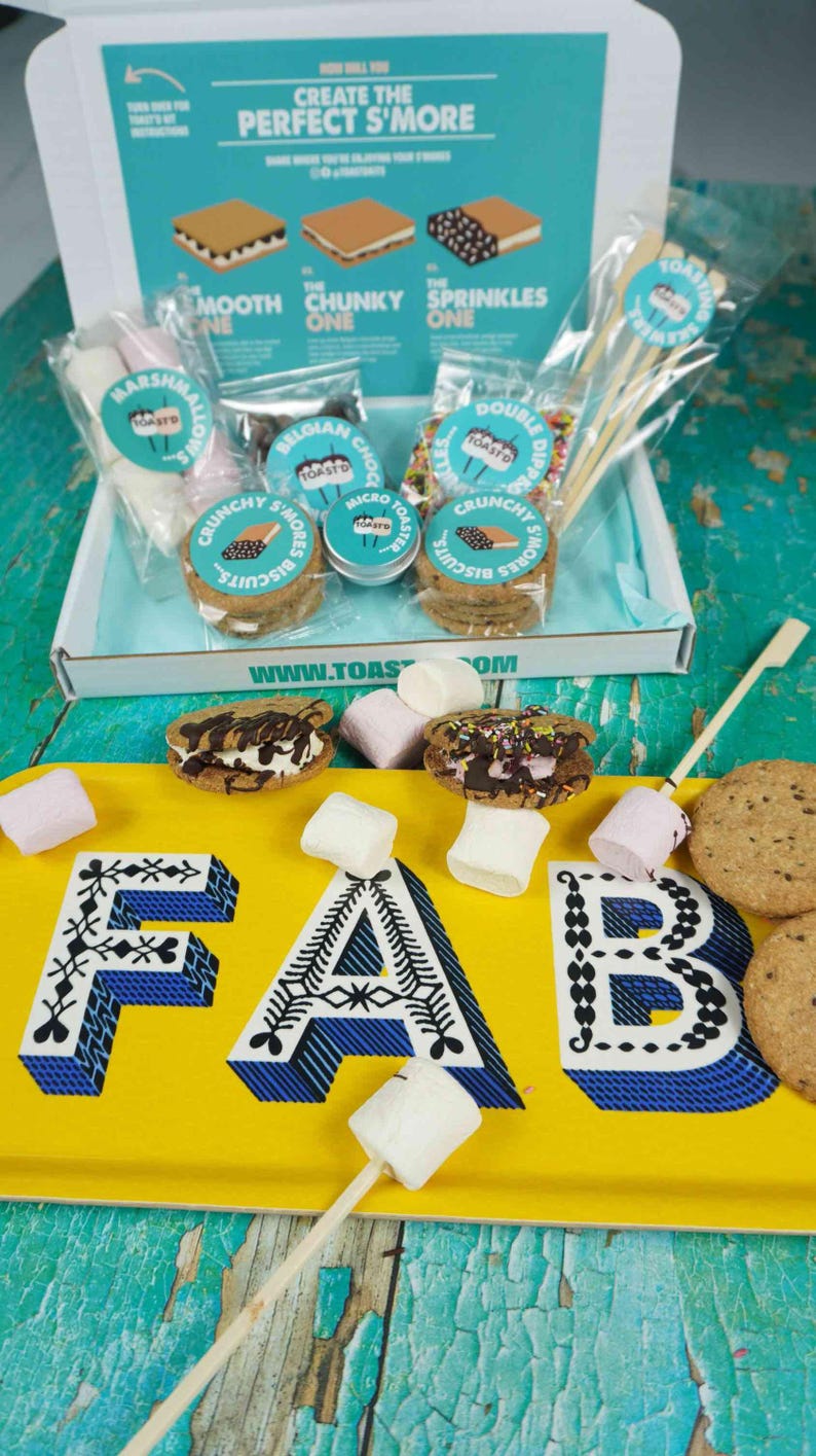 May include: A s'mores kit with ingredients and instructions. The kit includes marshmallows, chocolate, cookies, and skewers. The box has text that says "Create the Perfect S'More". A yellow tray with the word "FAB" is in the foreground.