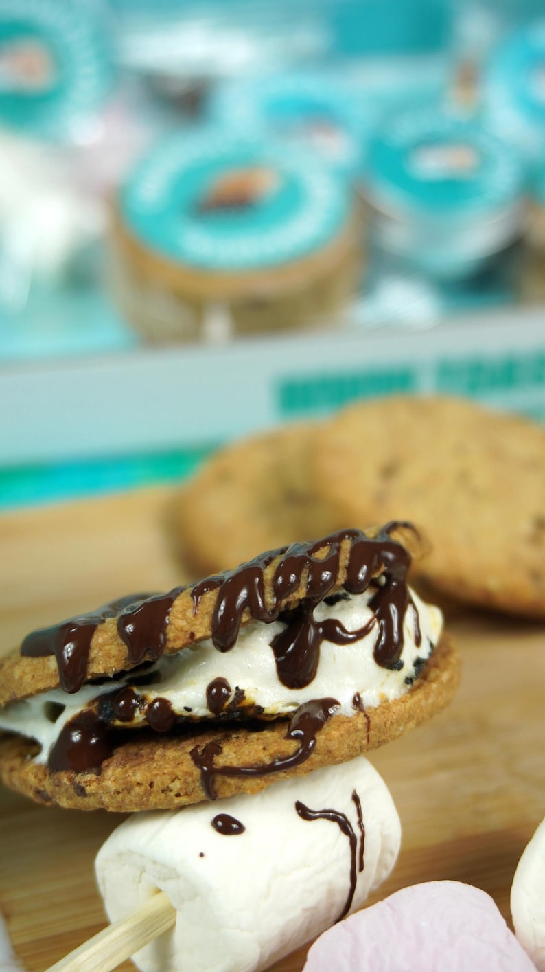 May include: A close-up of a s'more, featuring a toasted marshmallow and chocolate drizzle between two cookies. A marshmallow on a stick is in the foreground. The background shows more cookies and containers.