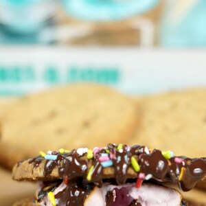 May include: A close-up of a s'more with chocolate drizzle and colorful sprinkles. The s'more is made with two graham crackers, a toasted marshmallow, and a layer of chocolate. The background is blurred, with a light blue and white color scheme.