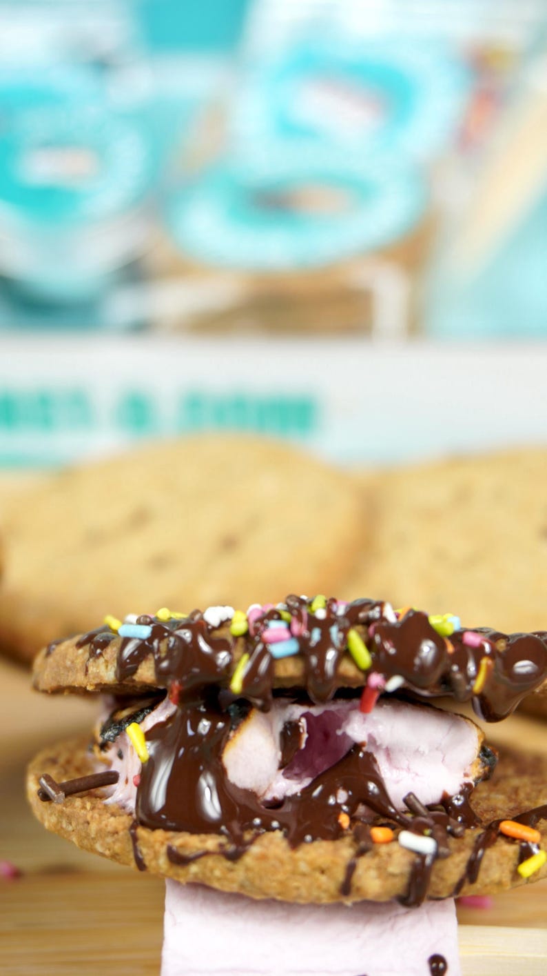 May include: A close-up of a s'more with chocolate drizzle and colorful sprinkles. The s'more is made with two graham crackers, a toasted marshmallow, and a layer of chocolate. The background is blurred, with a light blue and white color scheme.