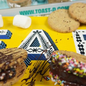 May include: A yellow tray with the text "www.toast-d.com" printed on it. The tray has two chocolate-covered cookies with sprinkles, a chocolate-covered cookie with nuts, and three white marshmallows. The tray is on a blue and green wooden surface.