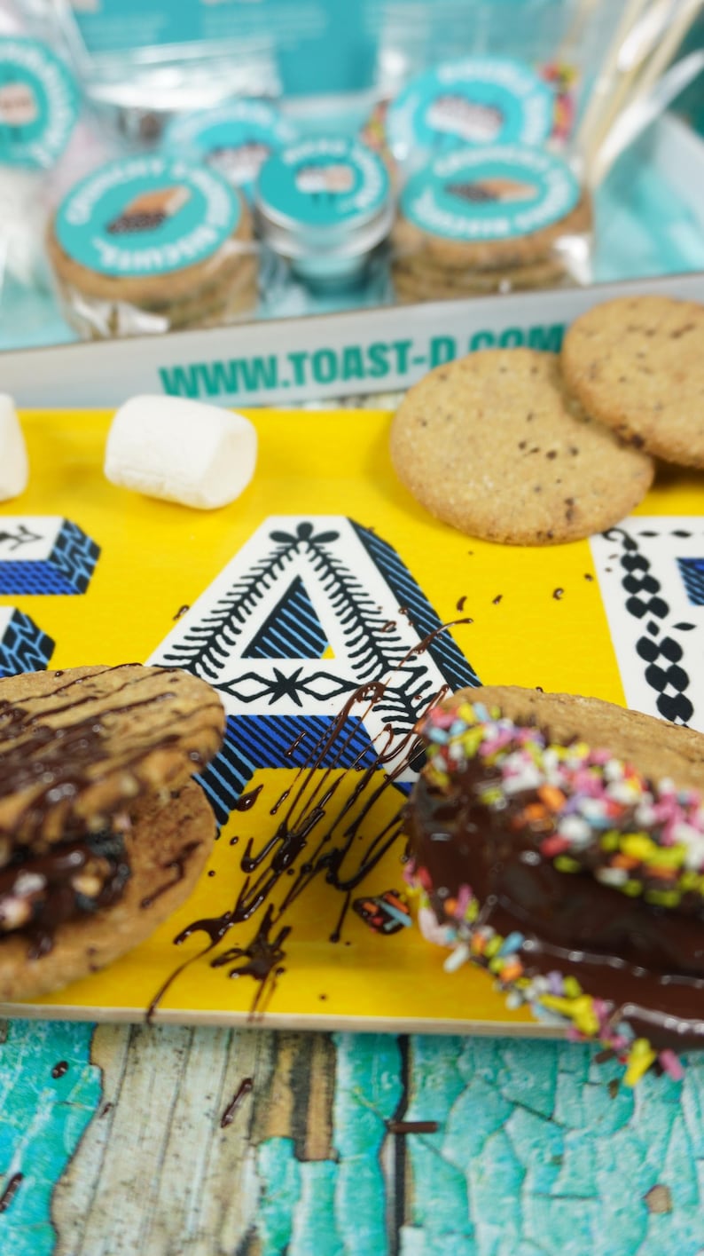 May include: A yellow tray with the text "www.toast-d.com" printed on it. The tray has two chocolate-covered cookies with sprinkles, a chocolate-covered cookie with nuts, and three white marshmallows. The tray is on a blue and green wooden surface.