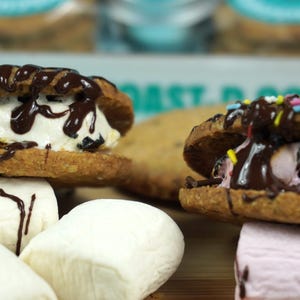 May include: Close-up of two s'mores, each made with two golden-brown cookies, marshmallow, and drizzled chocolate. One s'more has white marshmallow, the other has pink marshmallow and sprinkles. Several marshmallows are in the foreground.