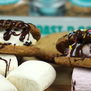 May include: Two chocolate chip cookie sandwiches with different fillings. One has vanilla ice cream and chocolate sauce, the other has pink marshmallow and chocolate sauce with sprinkles. Both are on sticks with marshmallows on the side.