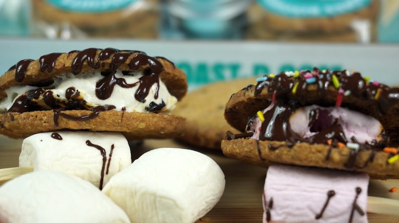 May include: Two chocolate chip cookie sandwiches with different fillings. One has vanilla ice cream and chocolate sauce, the other has pink marshmallow and chocolate sauce with sprinkles. Both are on sticks with marshmallows on the side.