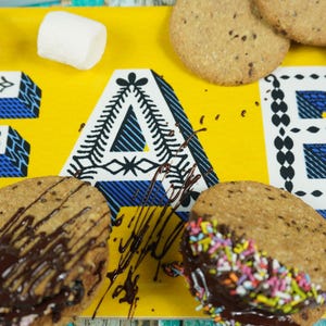 May include: A yellow tray with the word "FAB" written in black and white patterned letters. There are several cookies on the tray, some with chocolate and sprinkles. There are also marshmallows on the tray.