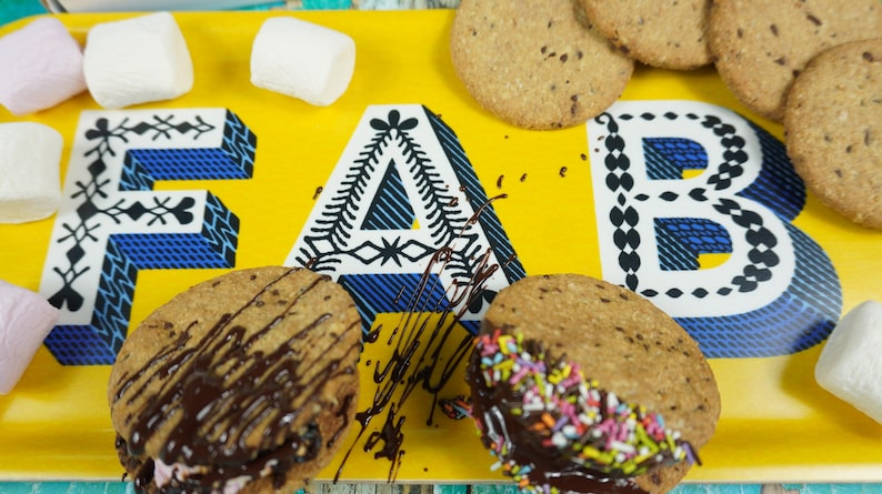 May include: A yellow tray with the word "FAB" written in black and white patterned letters. There are several cookies on the tray, some with chocolate and sprinkles. There are also marshmallows on the tray.