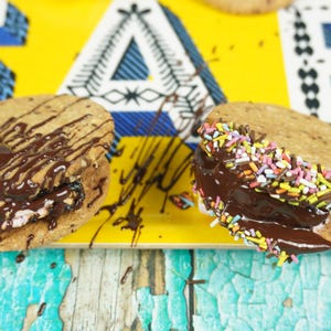 May include: Two chocolate-covered cookies with different toppings. One cookie has chocolate drizzle and the other has colorful sprinkles. Both cookies are on a yellow tray with blue and white patterned letters.