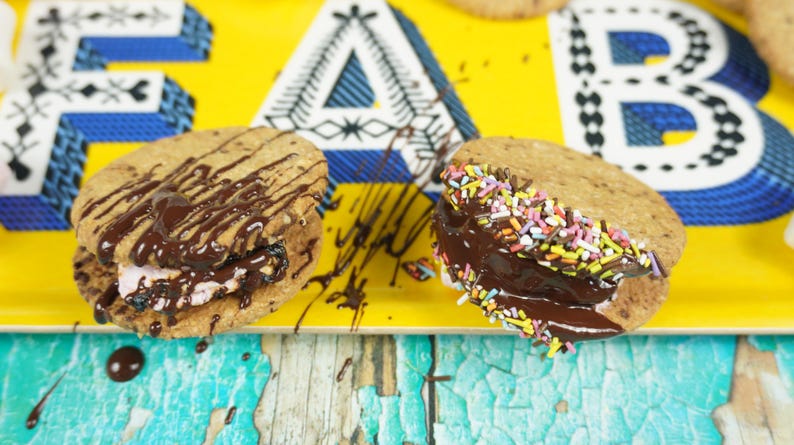 May include: Two chocolate-covered cookies with different toppings. One cookie has chocolate drizzle and the other has colorful sprinkles. Both cookies are on a yellow tray with blue and white patterned letters.