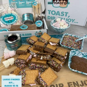 May include: A Toast'd s'mores kit featuring a white box with branding, a metal marshmallow roaster, biscuits, marshmallows, and chocolate. A wooden board displays assembled s'mores with chocolate and sprinkles, alongside bowls of toppings and a text card.