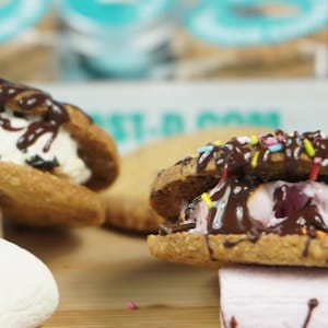 May include: Two chocolate-covered cookie sandwiches with different fillings, one with white ice cream and the other with pink marshmallow, on a wooden surface with colorful sprinkles.