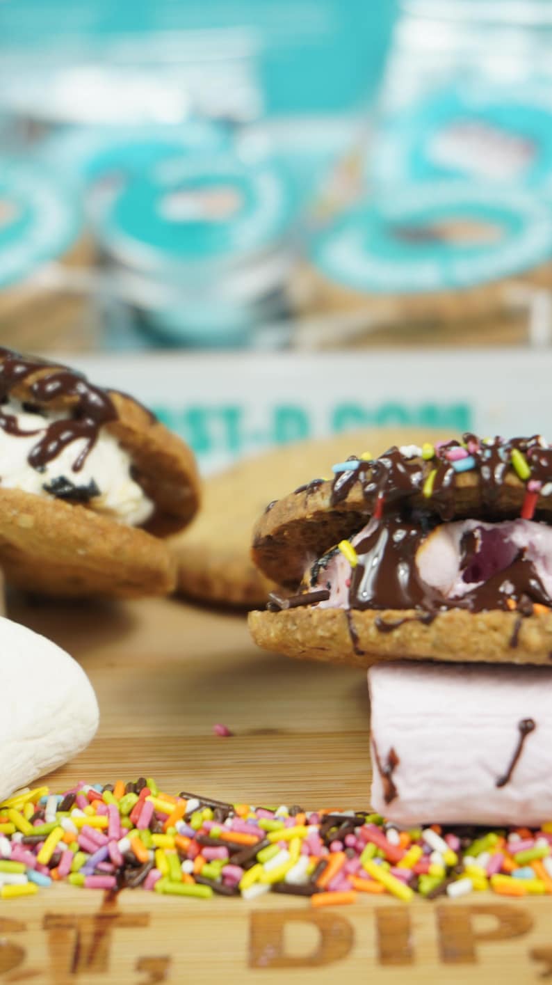 May include: Two chocolate-covered cookie sandwiches with different fillings, one with white ice cream and the other with pink marshmallow, on a wooden surface with colorful sprinkles.
