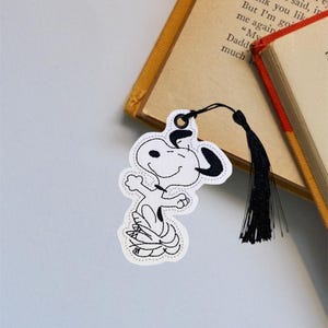 May include: A white and black embroidered bookmark featuring a cartoon dog with a black tassel. The bookmark is attached to a book with a yellow spine.