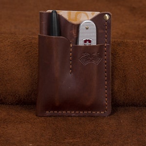 Puede incluir: Un organizador de bolsillo de cuero marrón con un bolígrafo y una navaja multiusos. El organizador tiene un borde cosido y un pequeño logotipo en la parte delantera.