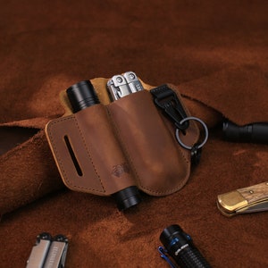 Puede incluir: Un portaherramientas multiusos de cuero marrón con dos bolsillos para llevar una linterna y una multiherramienta. El portaherramientas tiene un clip de metal y un llavero.