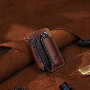 May include: A brown leather pocket organizer with a pen and a small folding knife. The organizer is on a brown suede surface.