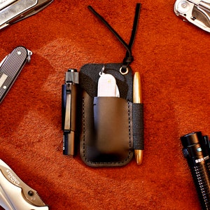Könnte beinhalten: Ein schwarzes Lederetui mit einem Riemen, das einen Stift, ein kleines Taschenmesser und eine Taschenlampe enthält. Das Etui liegt auf einer braunen Wildlederoberfläche.