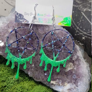 May include: A pair of green and black pentagram earrings with silver hooks. The earrings are dripping with green glitter. The earrings are on a background of purple and white crystals.