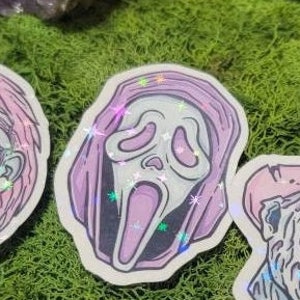 May include: A pink and white sticker with a ghost face mask. The mask has a wide open mouth and large eyes. The sticker has a holographic finish.