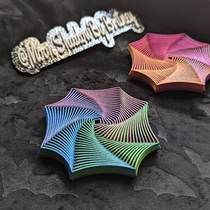 May include: Three colorful, geometric, spiral-shaped objects in shades of blue, green, pink, and orange. The objects are on a dark, textured surface. A silver-colored text that reads "@MoonShadowByBritney" is also visible.