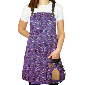 Può includere: Un grembiule con motivo viola e rosso con cinturini in pelle marrone e una tasca in pelle marrone. Il grembiule ha un motivo paisley.