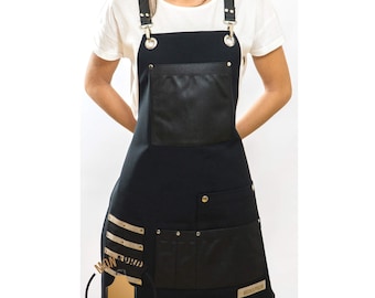 May include: Black canvas apron with leather accents and adjustable straps. The apron has multiple pockets, including a large front pocket and a smaller pocket on the side. The apron is branded with the word "MONDRIAN" in gold lettering.