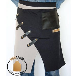 Puede incluir: Un delantal negro y beige con un bolsillo de cuero y correas ajustables. El delantal tiene un logotipo que dice "MONAPRONT Apron Art & Design".