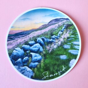 May include: Circular sticker with a landscape painting of a hillside, stone wall, and steps. The artwork shows blue rocks, green grass, and purple flowers under a sunset sky. The word "Simonside" is written in script.