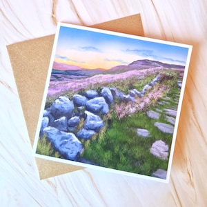 May include: A landscape art print depicting a stone wall, a path, and a field of purple flowers. The sky transitions from blue to orange and pink. The print is on a white card, with a brown envelope.
