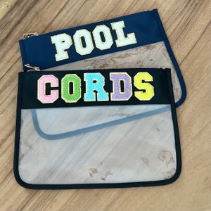 May include: Two clear plastic zippered pouches with black fabric borders. The top pouch has the word "POOL" in white letters with a gold outline. The bottom pouch has the word "CORDS" in colourful letters with a gold outline.