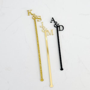 May include: Three personalized drink stirrers with initials. One is gold glitter, one is gold, and one is black. The initials are K&S, J&M, and A&D. These are ideal for weddings or other special events.