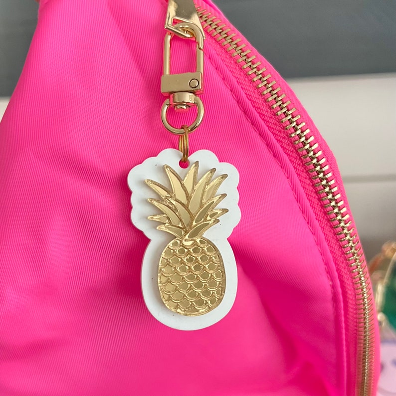 May include: A pink bag with a gold zipper and a gold pineapple keychain. The pineapple is on a white background.