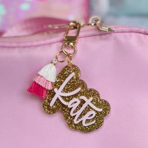 May include: A gold glitter keychain with the name "Kate" in white lettering and a pink and white tassel.