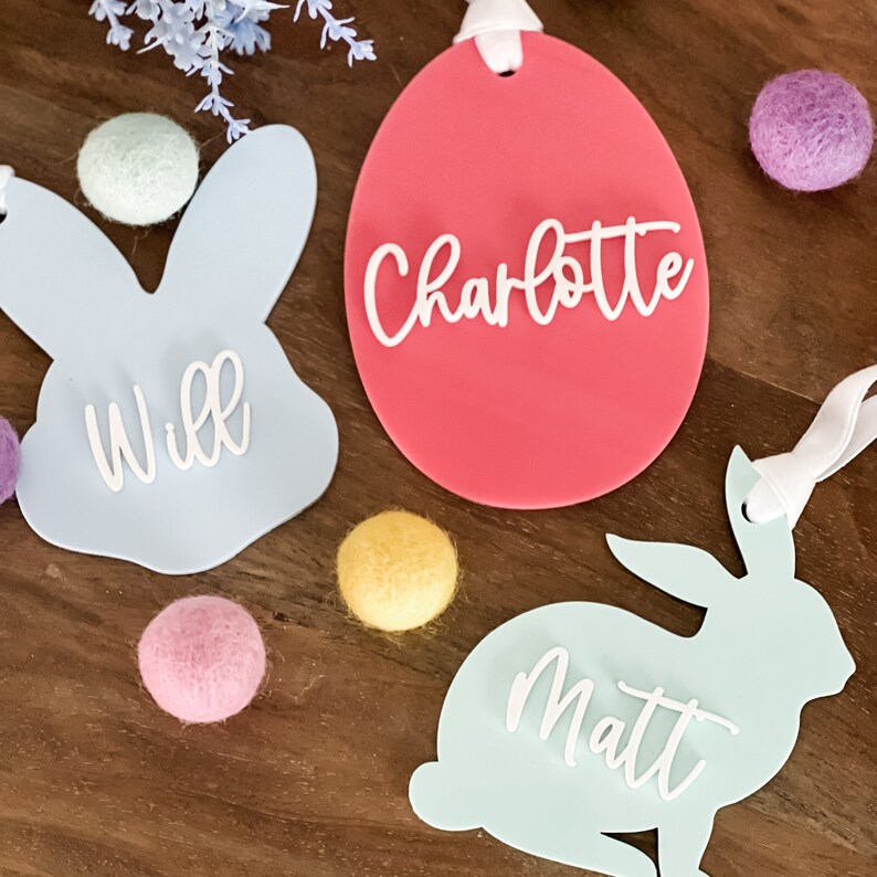 May include: Three personalized Easter decorations: a light blue bunny with the name "Will", a pink egg with the name "Charlotte", and a light blue bunny with the name "Matt".